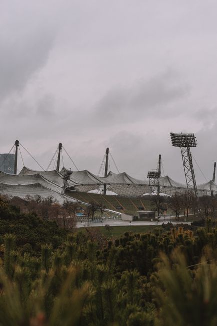The image depicts a large outdoor sports stadium with a distinctive tensile fabric roof supported by multiple high masts and cables, situated behind a foreground of green foliage and trees. The stadiu